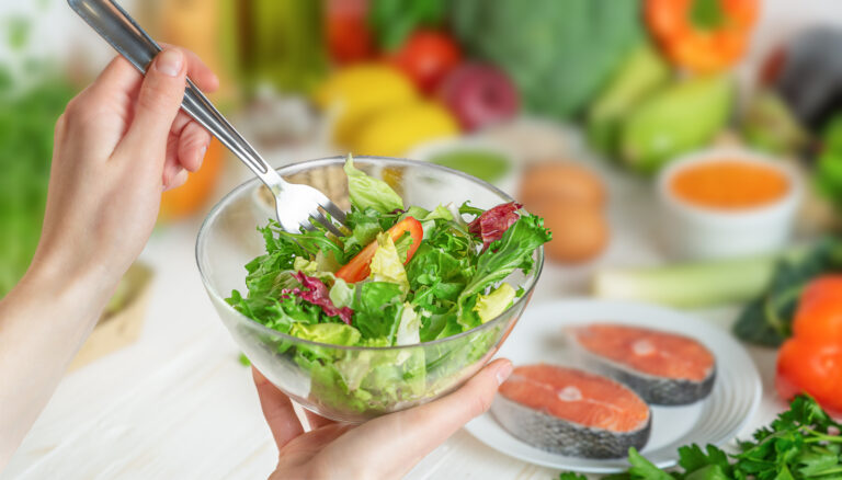 Image of a woman holding a salad bowl with fish and vegetables in the background representing healthy, anti-inflammatory foods