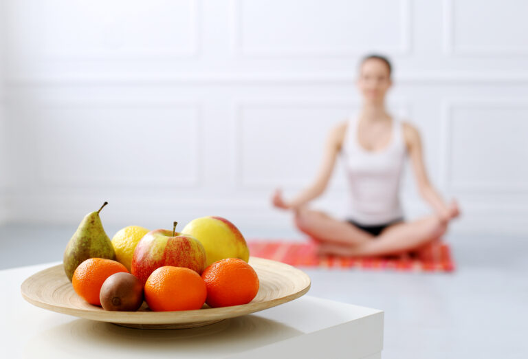 Plate of fresh fruit in the foreground and a woman doing yoga in the background representing mindfulness and health